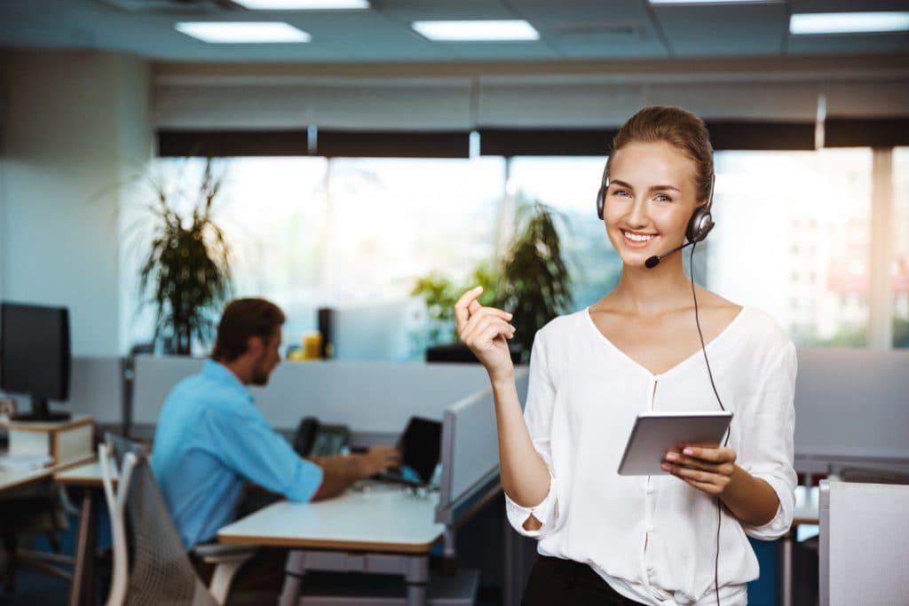 A smiling young woman wearing a headset and holding a tablet in a modern office setting, with a man working on a computer in the background.